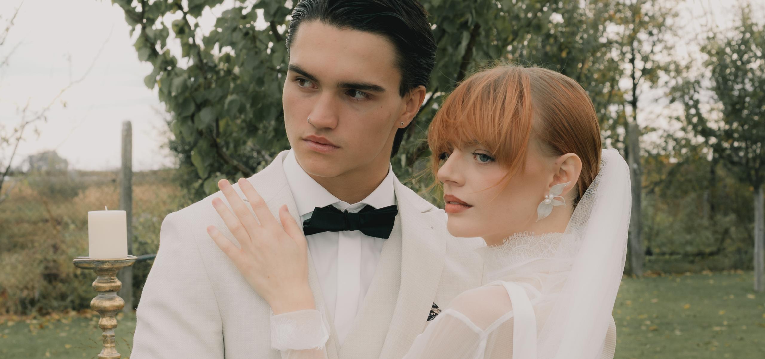 Nevesta s perlovými náušnicami stojí pri ženíchovi, ich pohľady smerujú mimo objektív, zachytení v exteriéri medzi stromami.