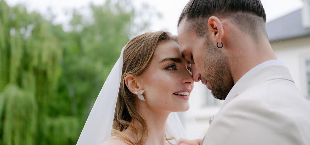 Nevesta so závojom a perlovými náušnicami stojí tvárou pri ženíchovi, obaja sú zachytení v jemnom romantickom momente v exteriéri.