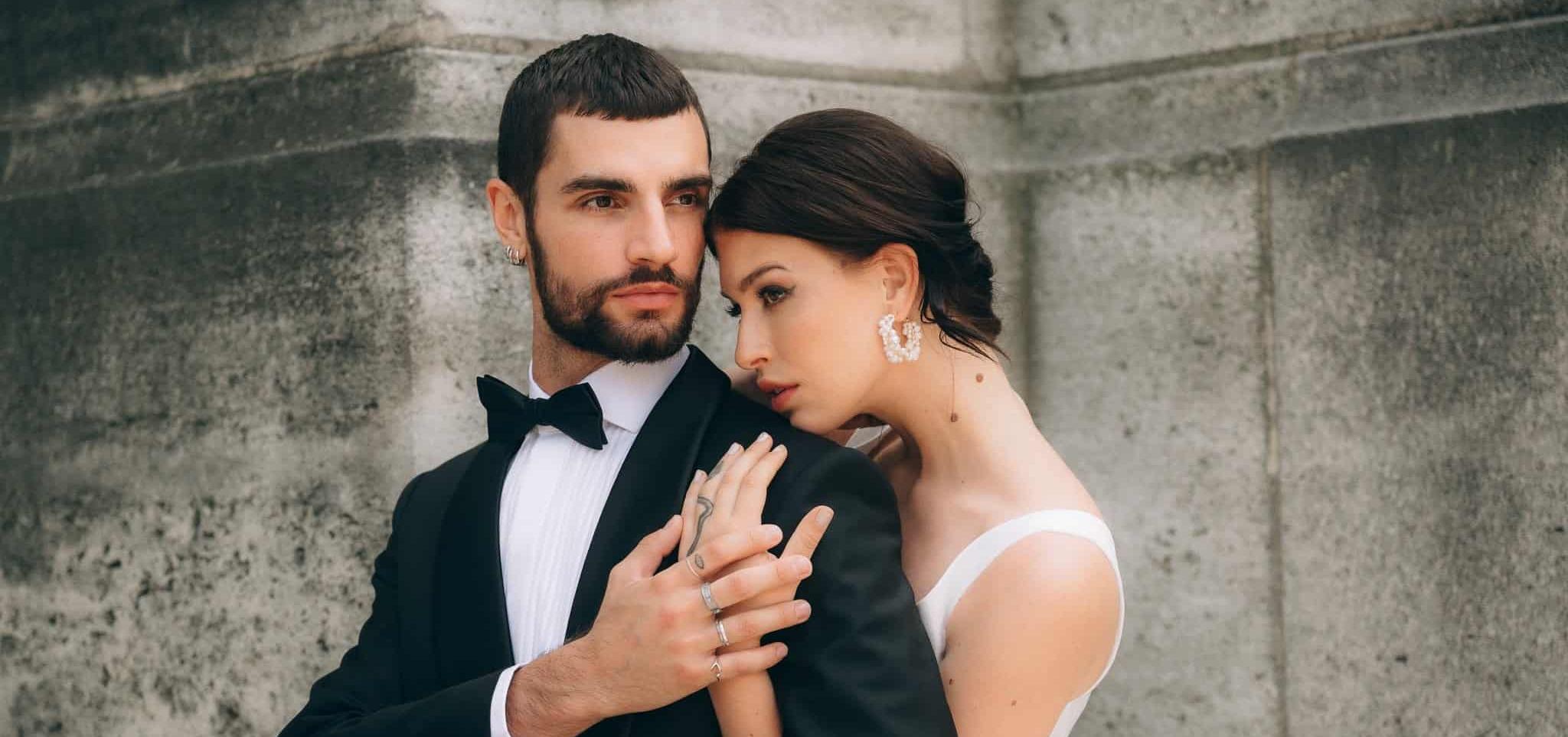 Nevesta s výraznými perlovými náušnicami stojí v objatí ženícha v smokingu, obaja sú zachytení pri kamennej stene v jemnom romantickom momente.