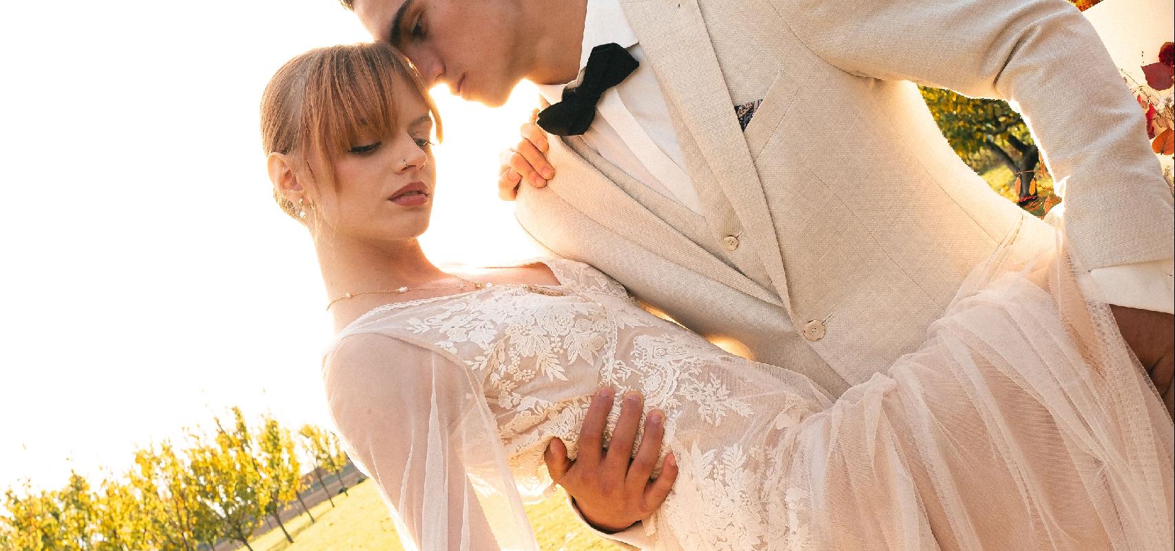 Nevesta s kvetinovými náušnicami stojí pri ženíchovi, ich pohľady smerujú mimo objektív, zachytení v exteriéri medzi stromami.