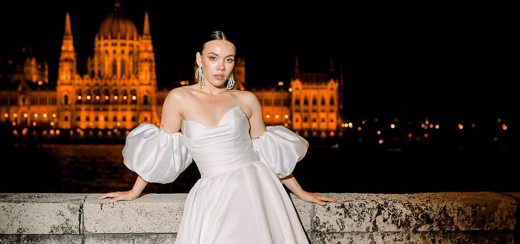 nevesta stojaca v dlhých šatách v budapešti, v pozadi stoje monumentálna osvetlená budova