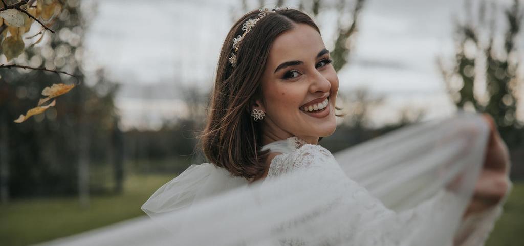 Nevesta s kvetinovými náušnicami stojí pri ženíchovi, ich pohľady smerujú mimo objektív, zachytení v exteriéri medzi stromami.