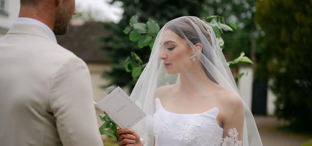 Nevesta so závojom a perlovými náušnicami stojí tvárou pri ženíchovi, obaja sú zachytení v jemnom romantickom momente v exteriéri.