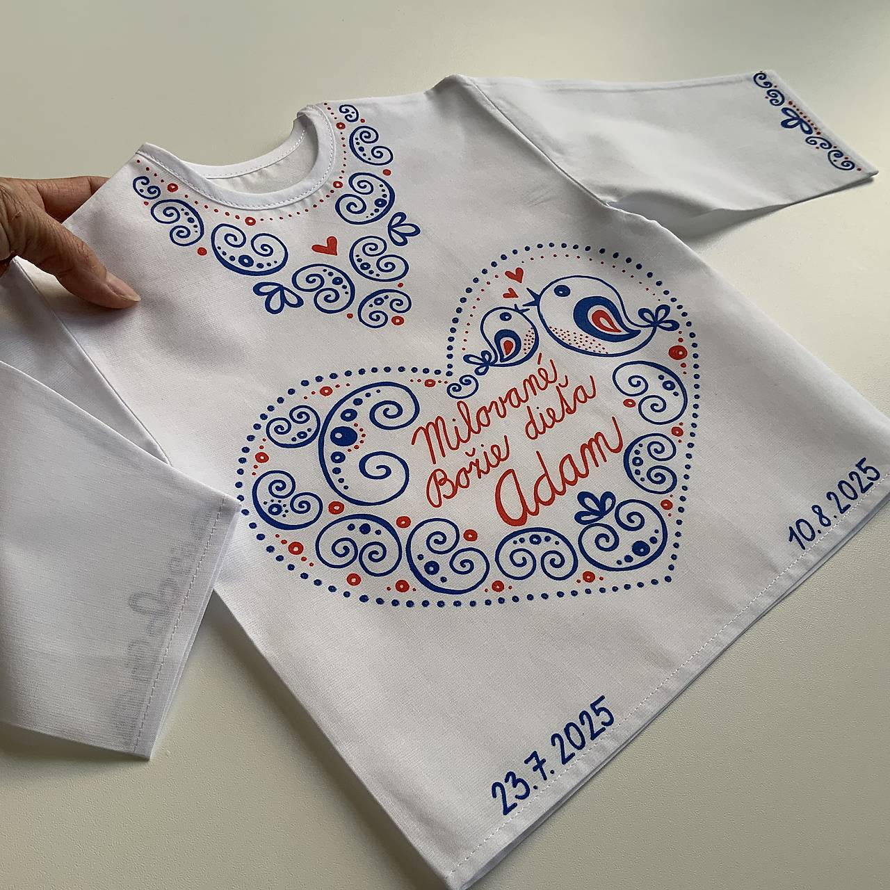 Krstná košieľka maľovaná s autorským ludovoladeným motívom v srdiečku aj okolo goliera a rukávov v modro-červenej 