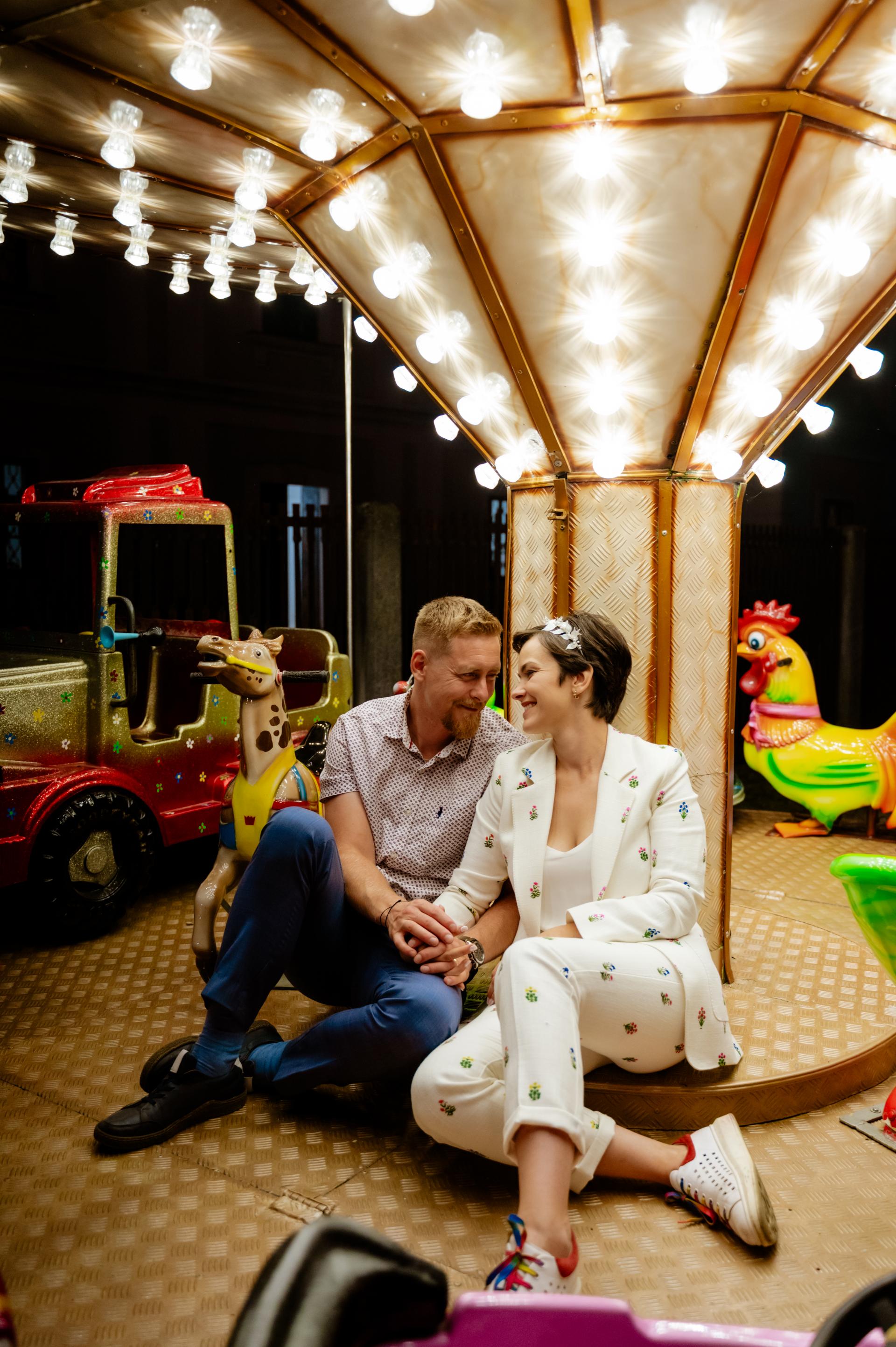 nevesta a ženích sediaci pri rozsvietenom kolotoči v lunaparku. Nevesta má biely vzorovaný kostým a na hlave bielu čelenku s lístkami.