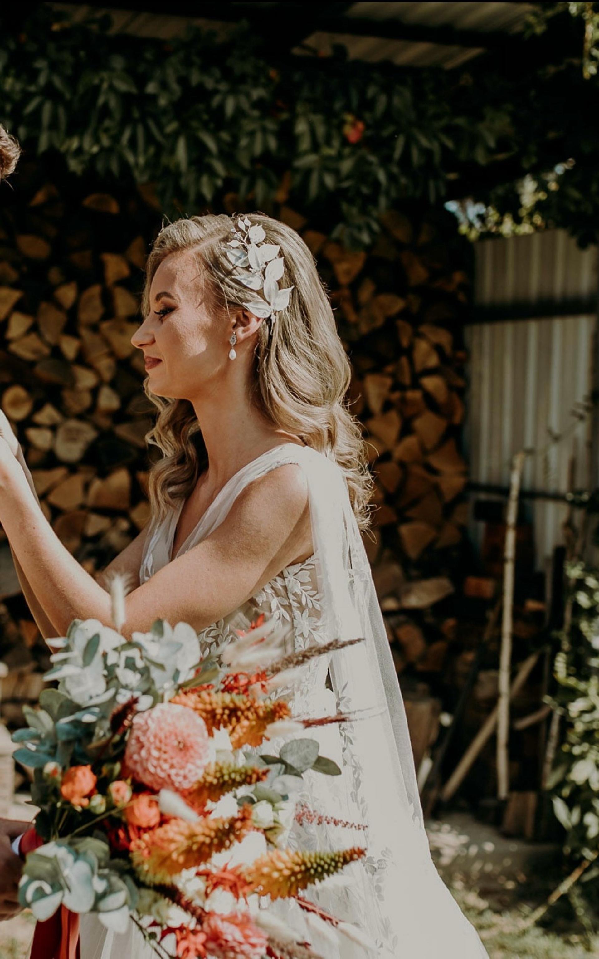 Nevesta v svadobných šatách s jemnou vlasovou ozdobou a perlovými náušnicami, svadobný účes a kytica v prírodnom štýle