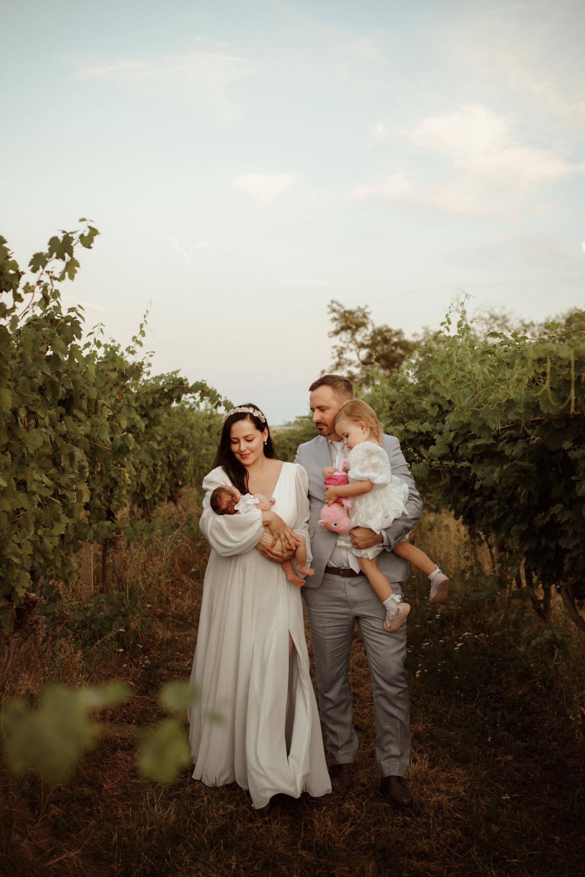 Rodinná fotografia vo vinici – mama v bielych šatách drží novorodenca, otec v svetlom obleku nesie malé dievčatko, spolu kráčajú medzi radmi viniča.