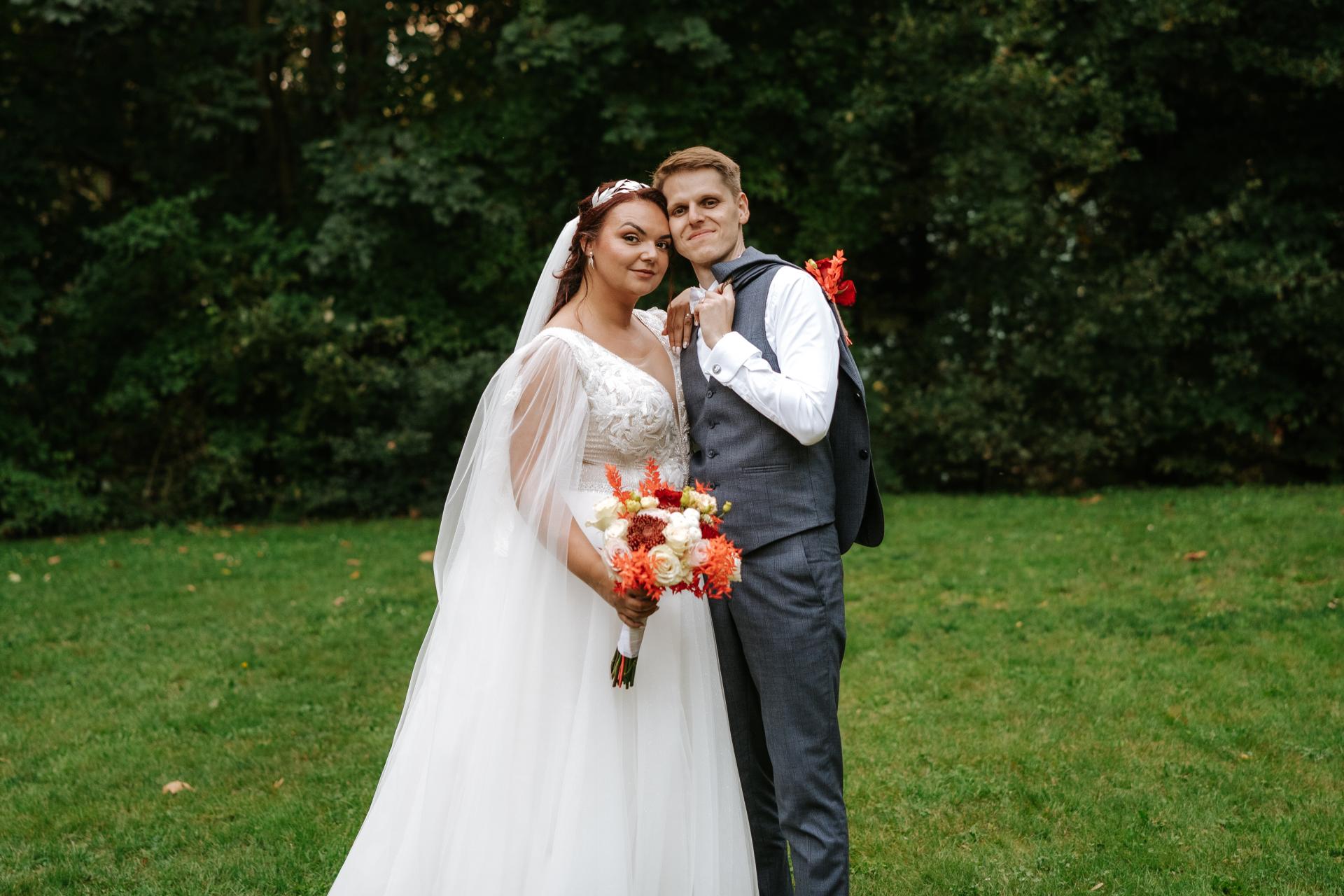 Nevesta v bielych svadobných šatách s kyticou a ženích v sivom obleku pózujú na tráve pred zeleným lesným pozadím.