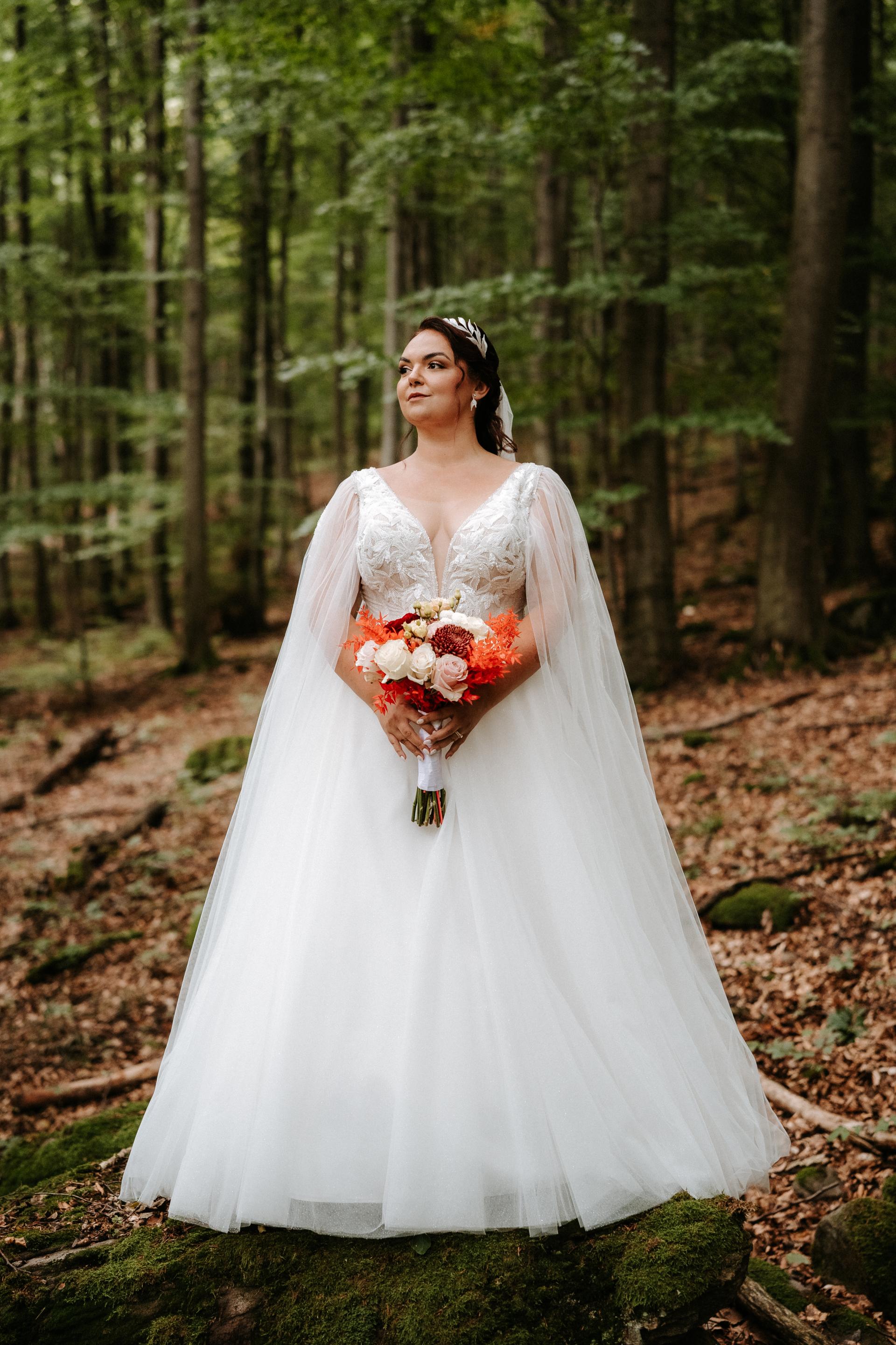 Nevesta v dlhých bielych svadobných šatách a so závojom stojí v lese na kameni, v ruke drží svadobnú kyticu v teplých odtieňoch.