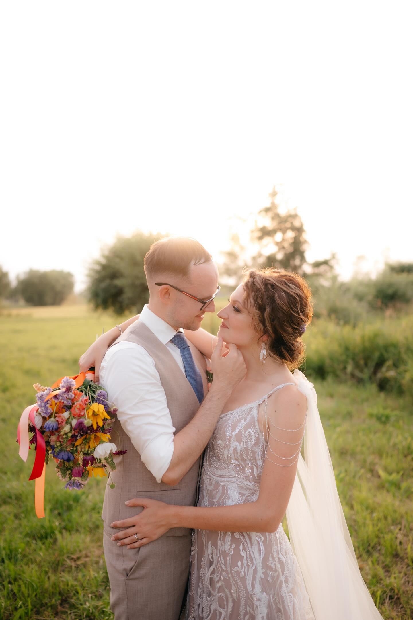 Nevesta a ženích objatí na lúke pri západe slnka, nevesta má zapletený účes, závoj a jemné svadobné šaty, ženích drží farebnú svadobnú kyticu.