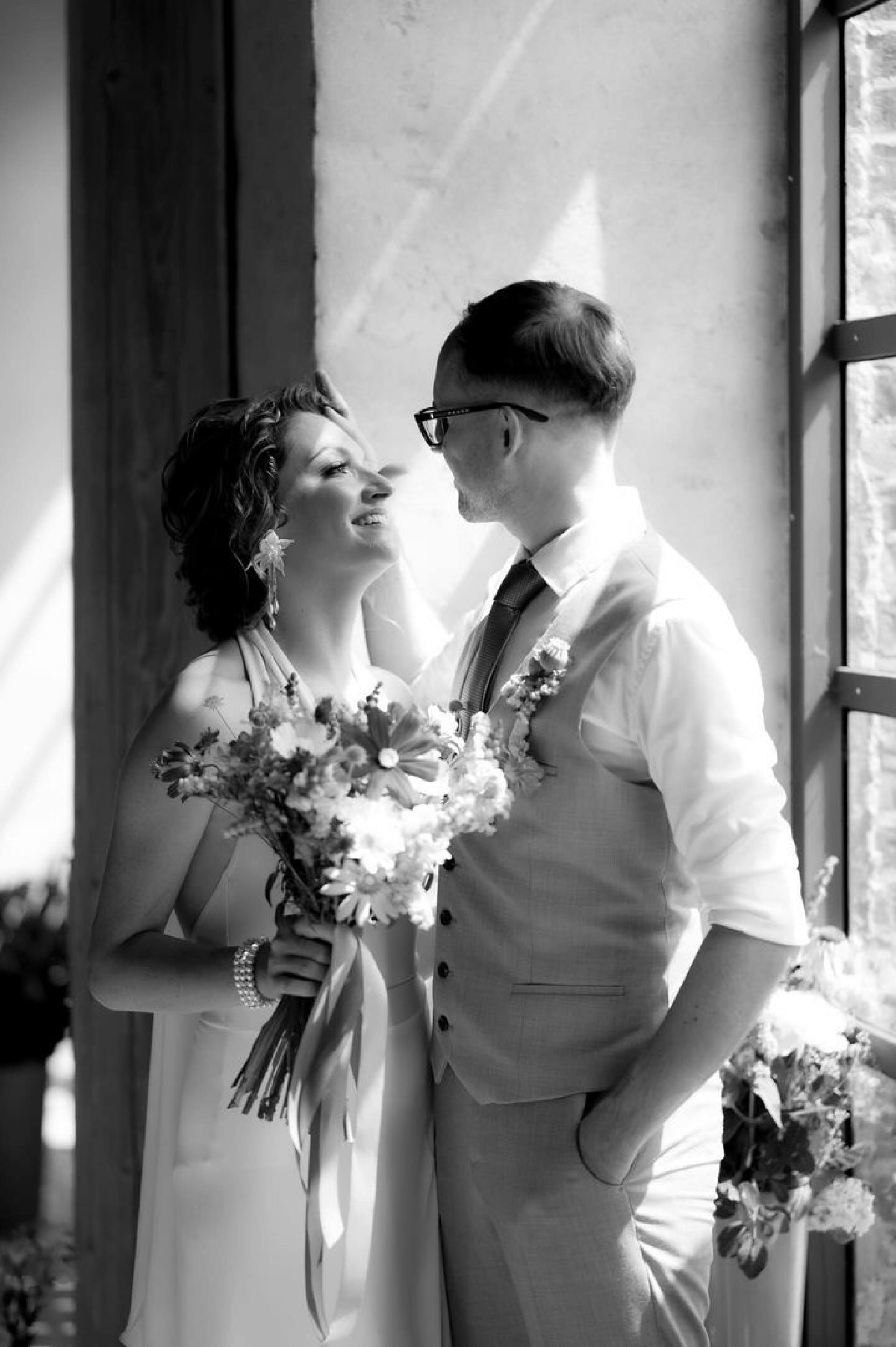 Čiernobiela svadobná fotografia nevesty a ženícha pri okne, pozerajú sa na seba, nevesta drží kyticu a má výrazné náušnice.