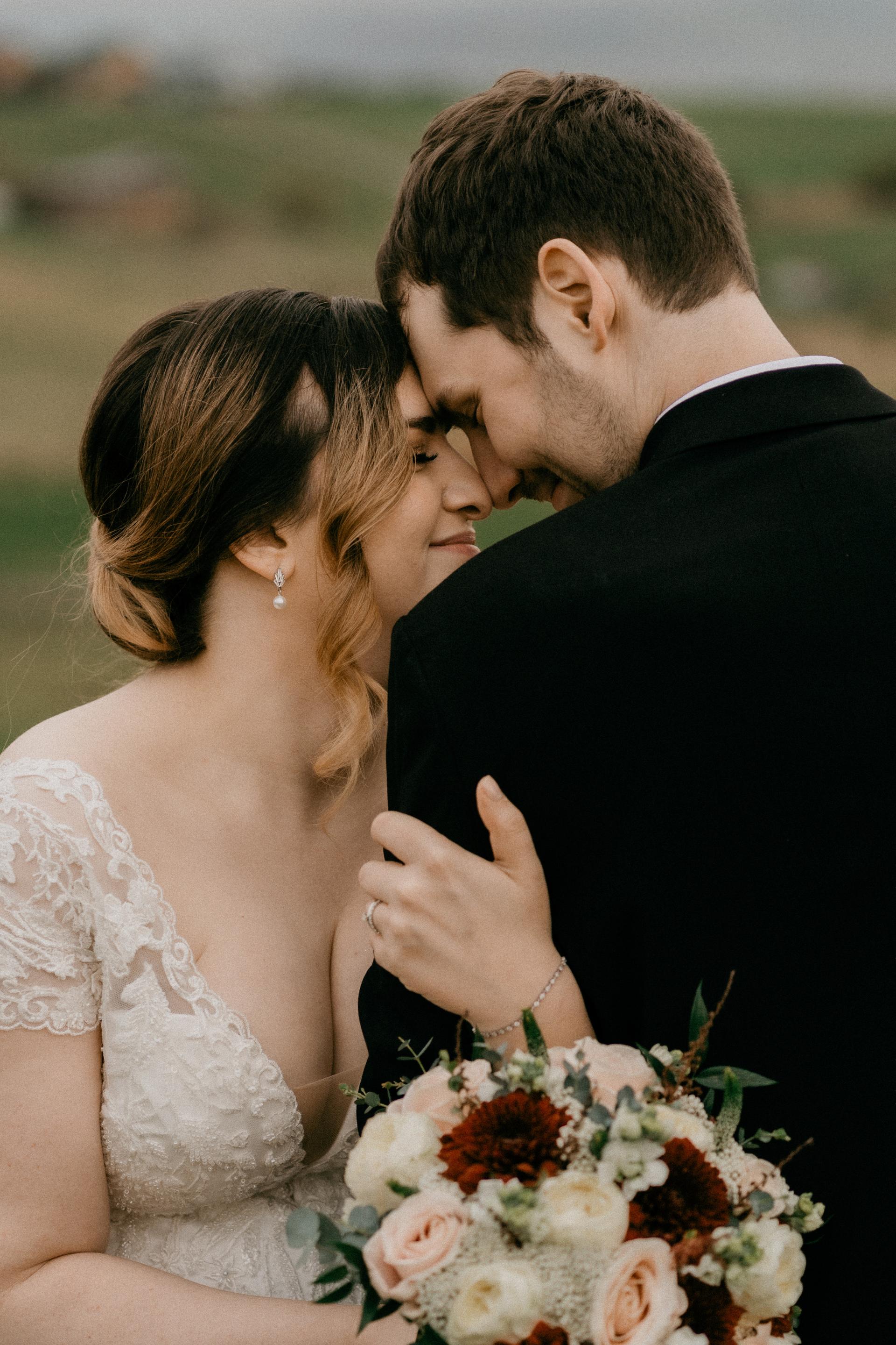 Nevesta v čipkovaných svadobných šatách sa nežne opiera o ženícha, v ruke drží svadobnú kyticu v krémových a hnedých tónoch, detailný záber v prírode.