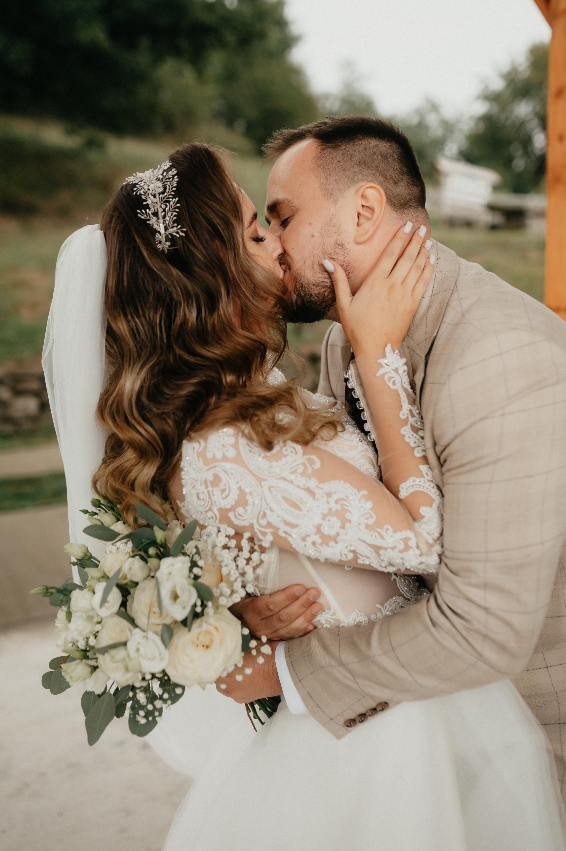 Nevesta a ženích sa bozkávajú, detailný záber na čipkované svadobné šaty nevesty, závoj a svadobnú kyticu z bielych ruží.