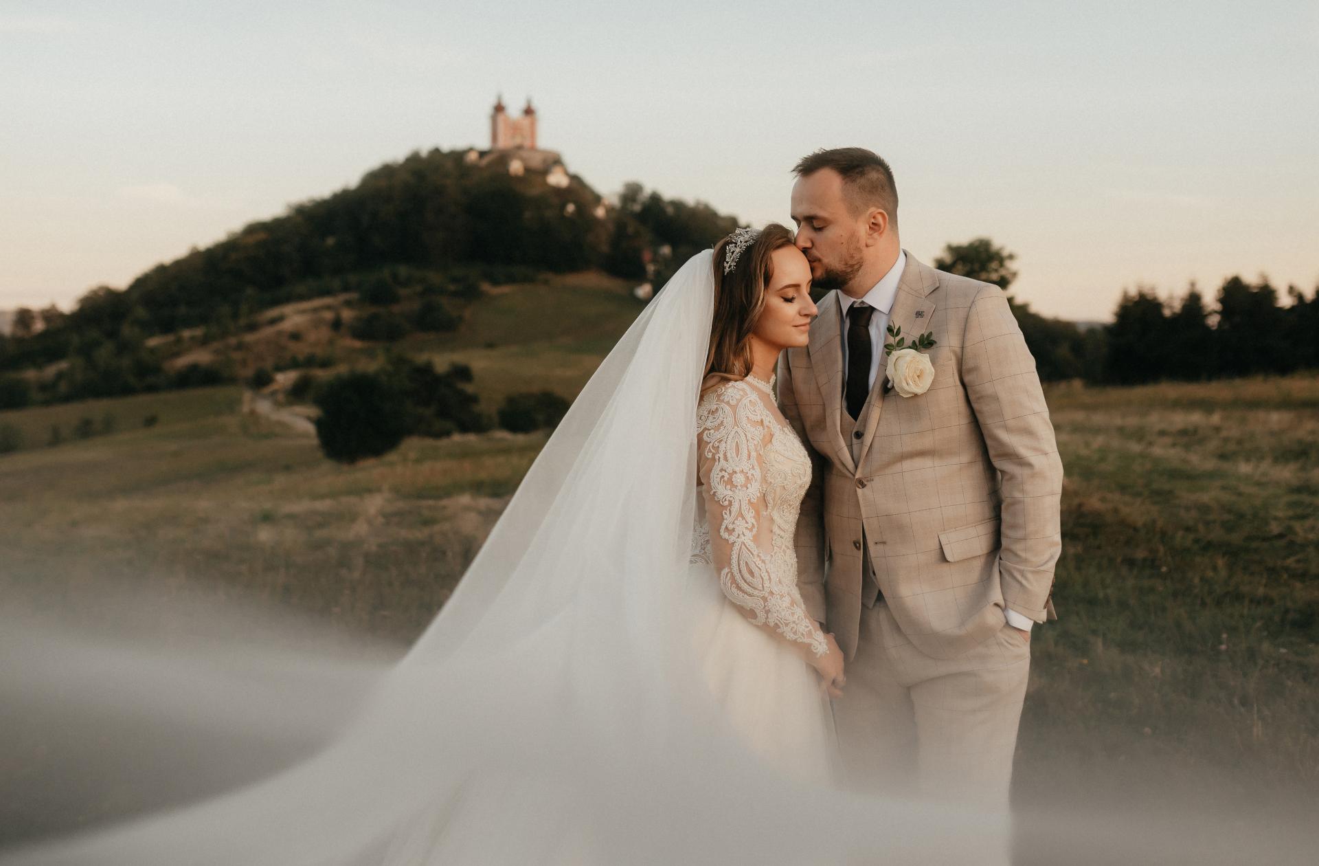 Nevesta so závojom a ženích v béžovom obleku stoja v krajine pri západe slnka, ženích bozkáva nevestu na čelo, v pozadí kopec s kostolíkom. banská štiavnica kalvárai