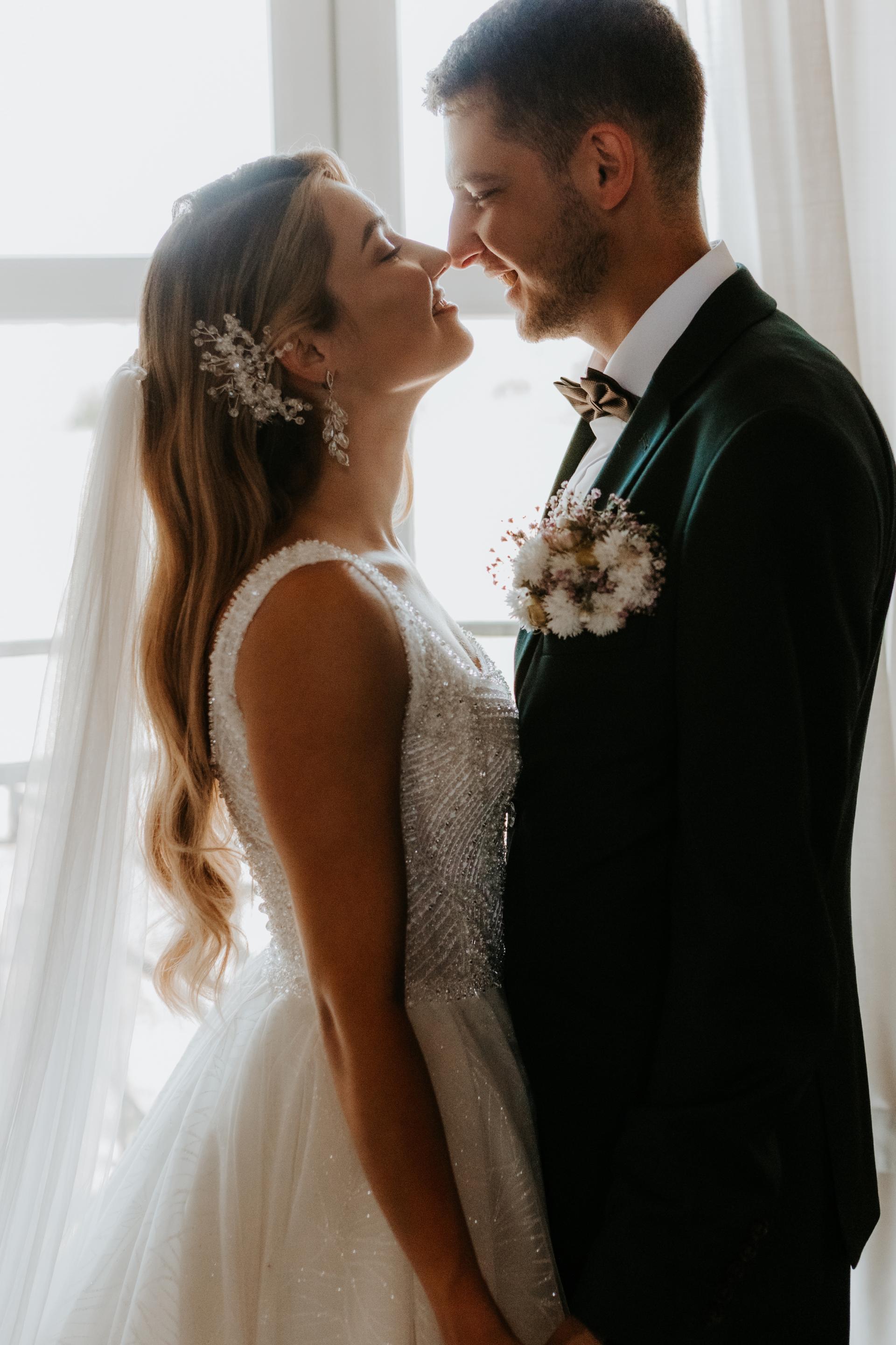 Nevesta a ženích stoja oproti sebe pri okne, usmievajú sa, nevesta má dlhé vlasy so závojom, výrazné svadobné náušnice a jemnú vlasovú ozdobu.