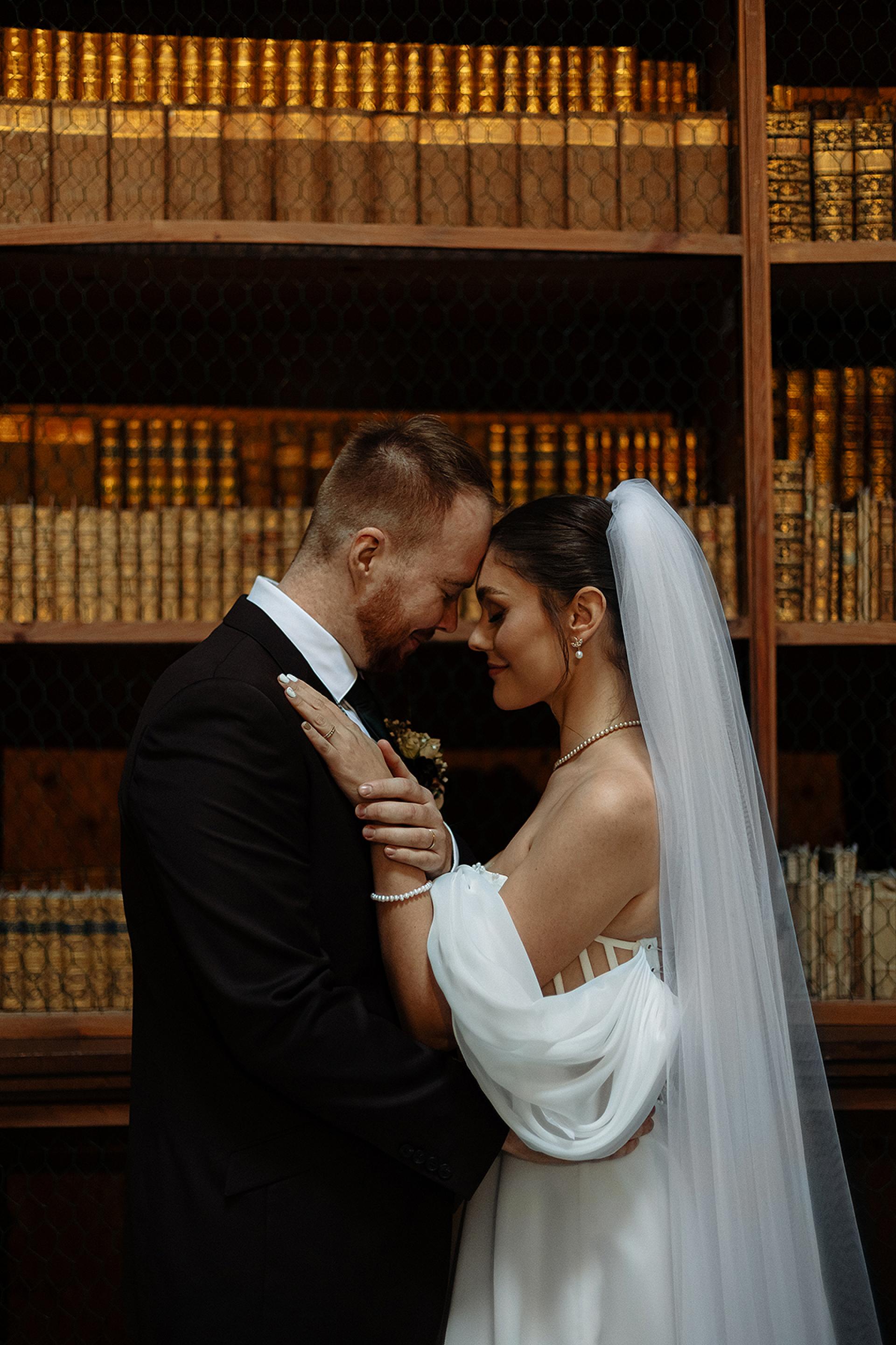 Nevesta so závojom a perlovými šperkami sa nežne dotýka čelom so ženíchom v knižnici, intímny portrét pred historickými knižnými policami.