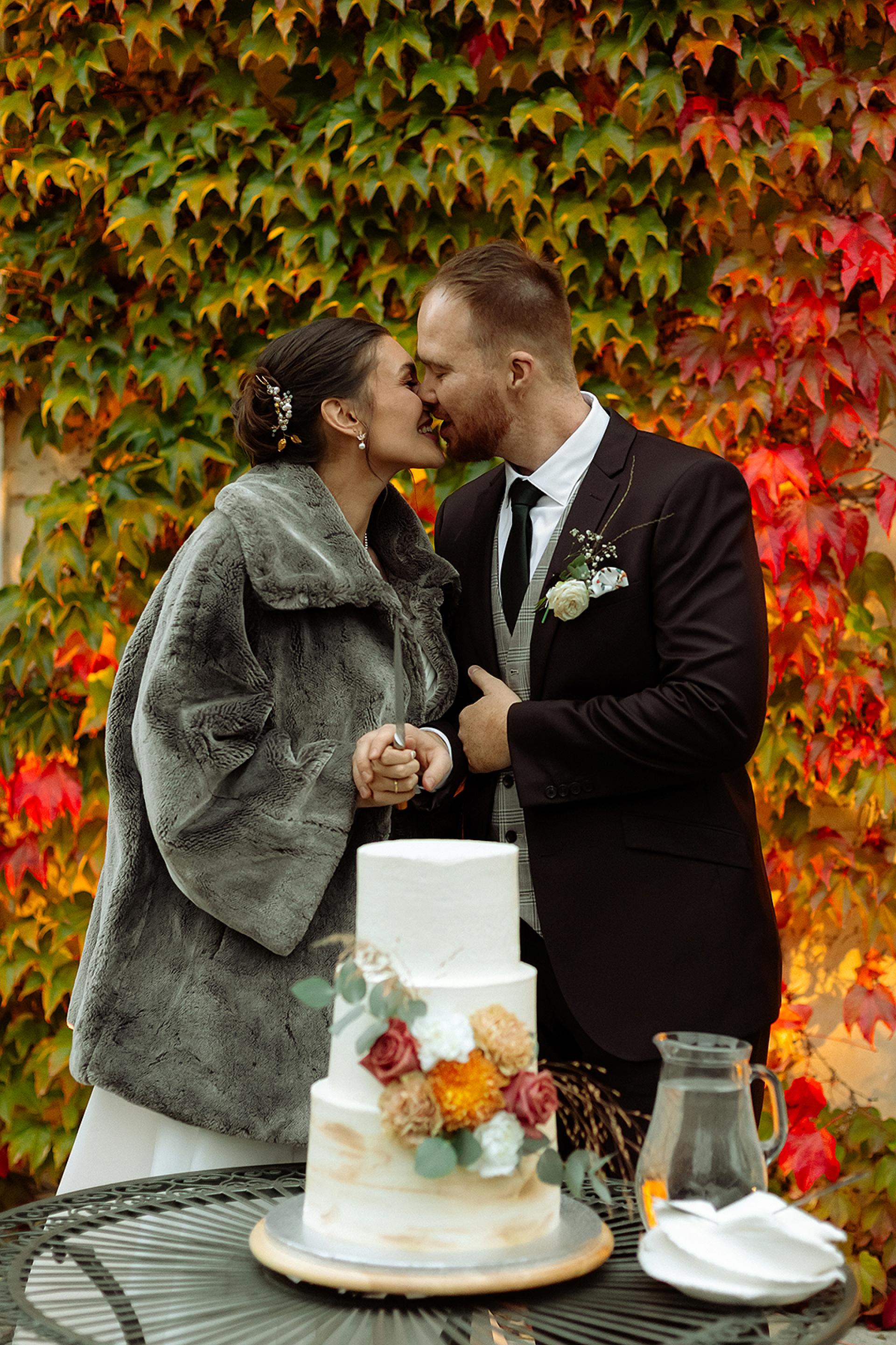 Nevesta a ženích sa bozkávajú pri krájaní svadobnej torty, jesenné farby lístia v pozadí, detail svadobnej oslavy vonku.