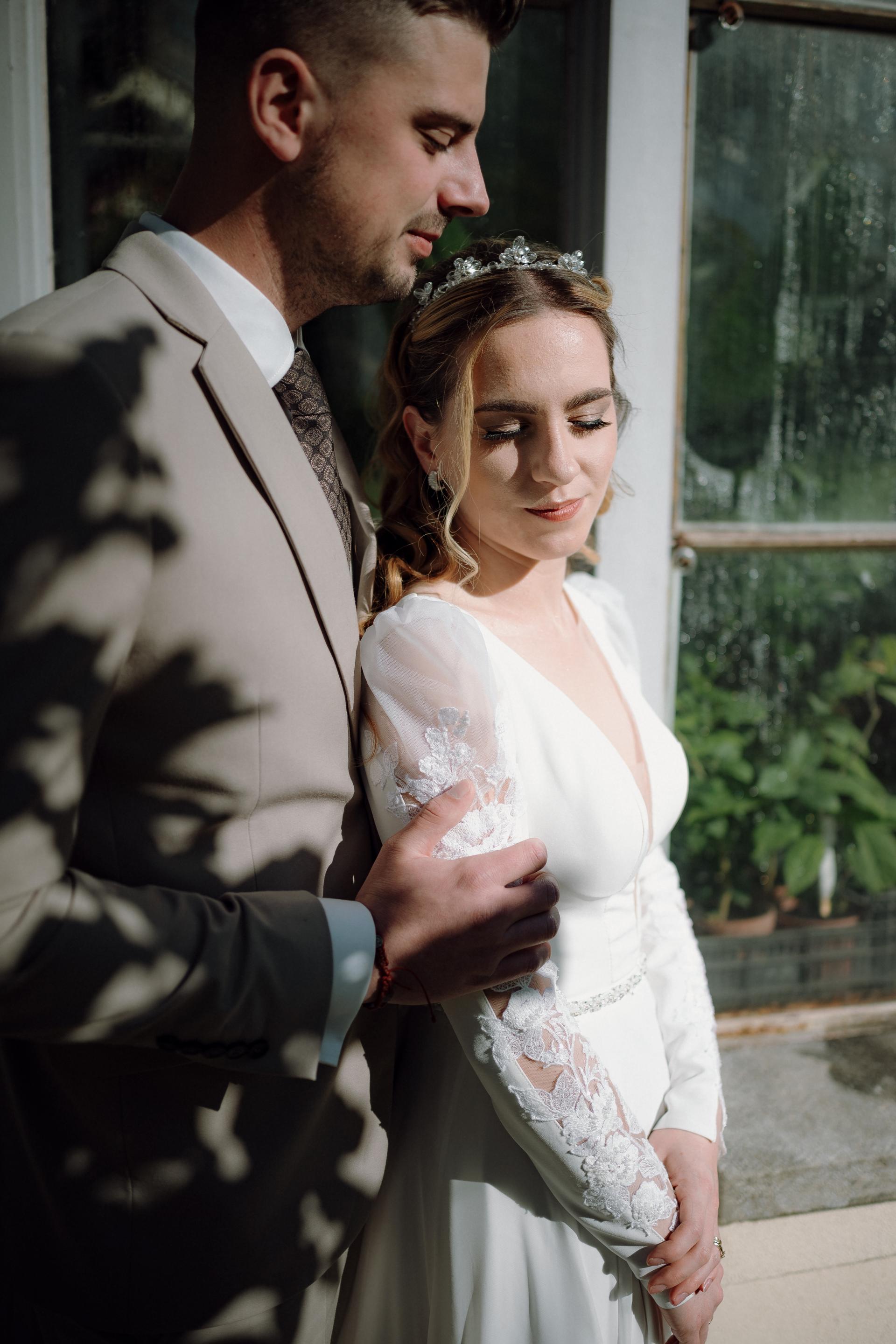 Nevesta v dlhých bielych svadobných šatách s čipkovanými rukávmi stojí pri okne, ženích v svetlom obleku ju jemne objíma zozadu, prirodzené svetlo a intímna atmosféra.