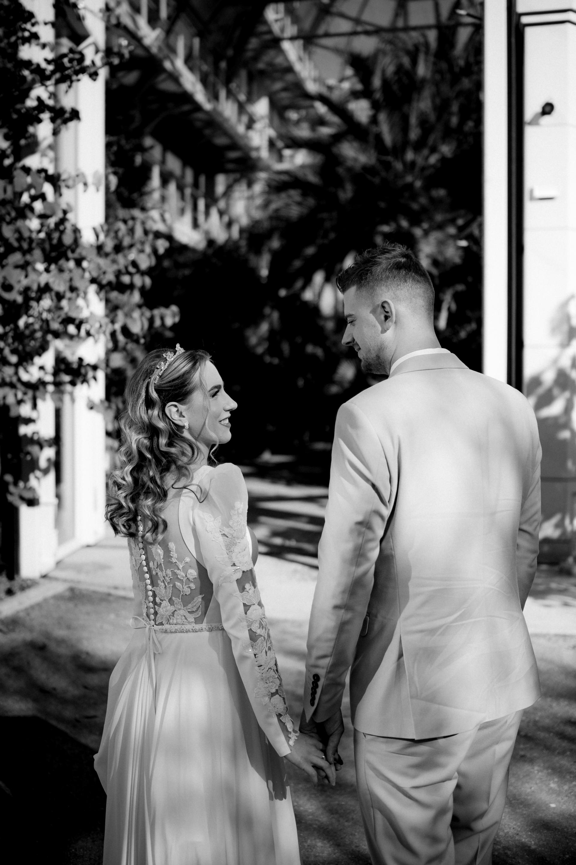 Čiernobiela svadobná fotografia nevesty a ženícha kráčajúcich ruka v ruke v exteriéri, nevesta má dlhé šaty s čipkovaným chrbtom a zapínaním na gombíky.